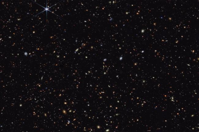 Distant deep field image showing hundreds of galaxies against the black of the night sky. The galaxies appear as faint white, blue, orange and red blobs. 
